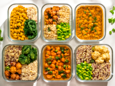 Zeven glazen bakjes gevuld met kleurrijke vegan meal prep maaltijden op een marmeren ondergrond